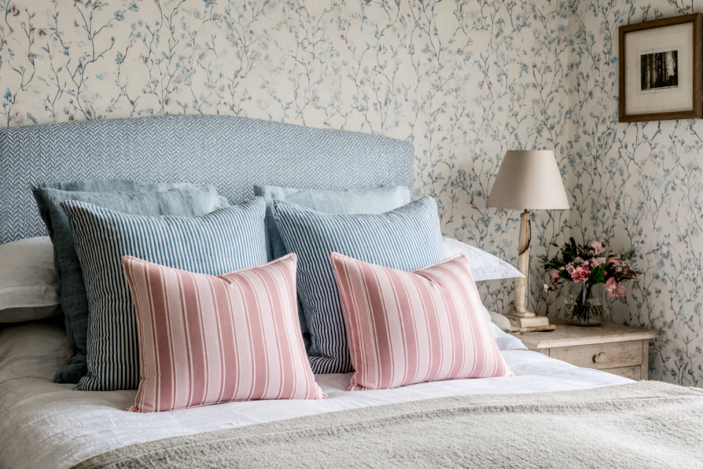Serene bedroom showing trailing floral wallpaper, pale blue herringbone headboard, and pink ticking stripe cushions