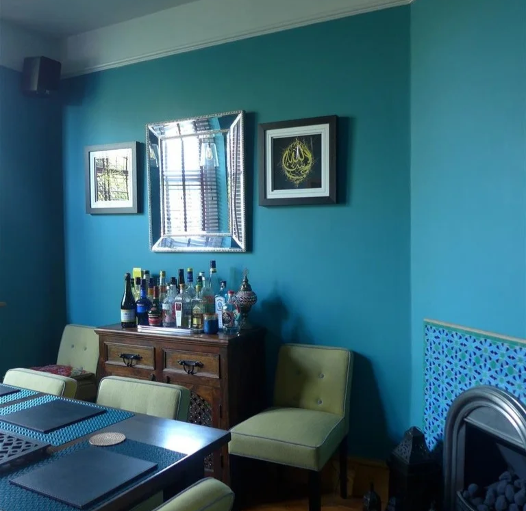 Dining room with teal walls and dim morning light, featuring soft green chairs, a dark wood sideboard with assorted bottles, and a fireplace with patterned blue tiles, illustrating how limited natural light in darker, north-facing rooms affects wall colour appearance.