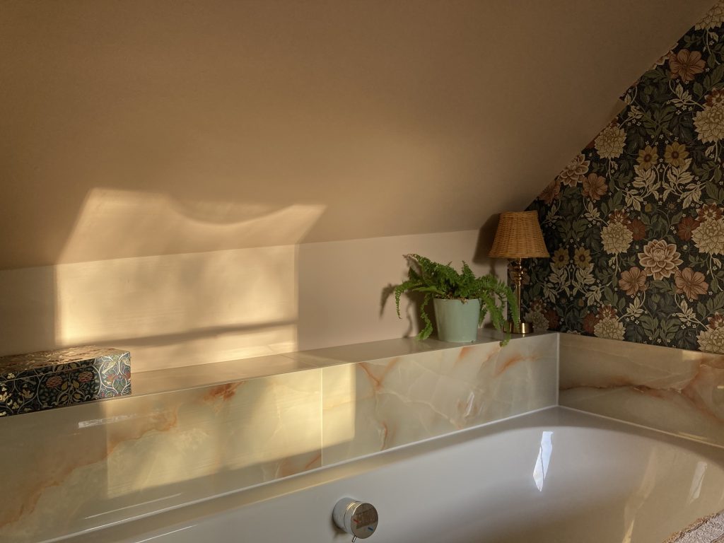 Soft evening light casts a warm glow on a blush-toned bathroom wall above a marble-trimmed bathtub, with a potted fern, wicker-shaded lamp, and floral wallpaper creating a cozy, vintage-inspired corner.