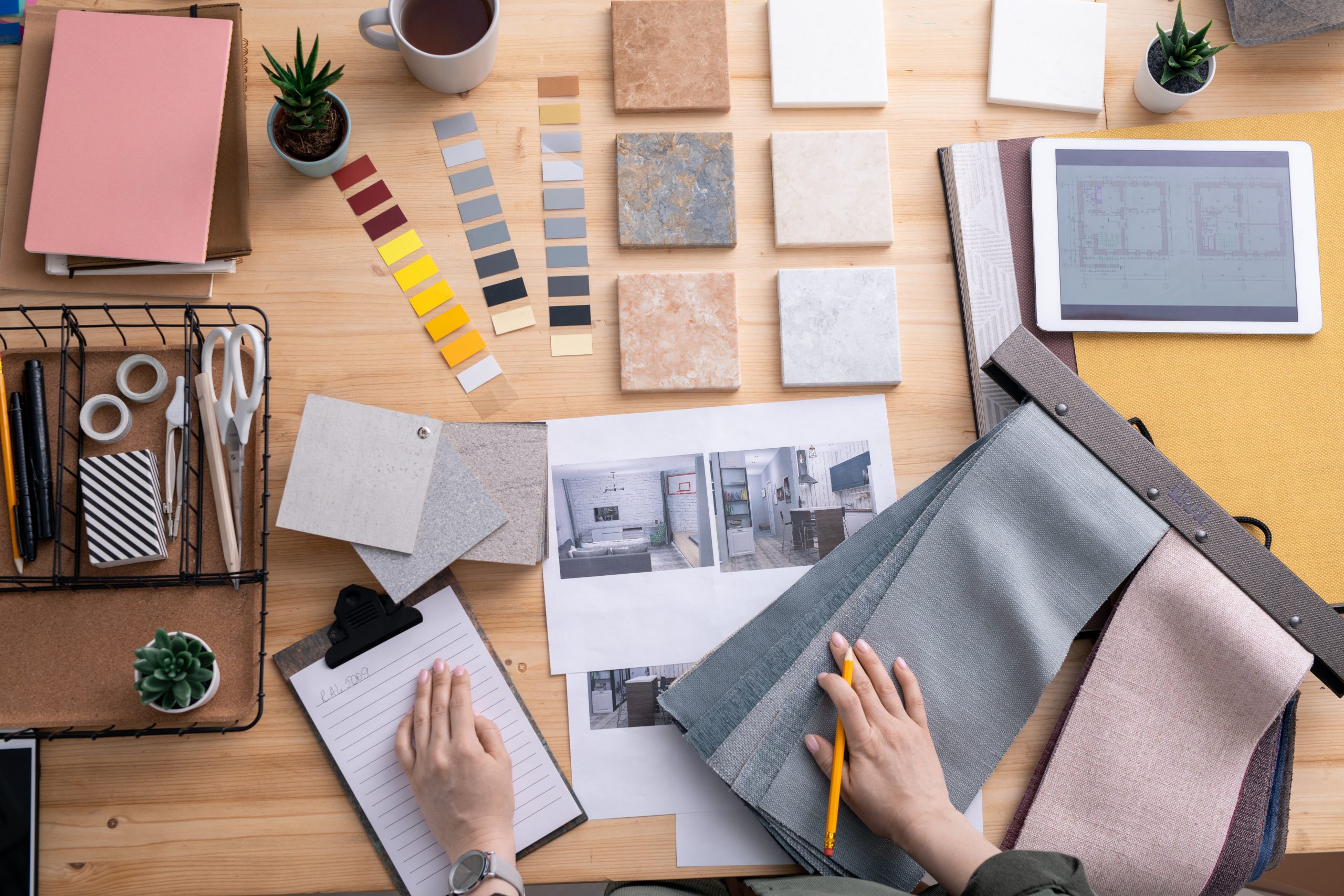Overhead photograph of an interior designer's desk, showing fabric and tile samples, colour charts, and technical drawings on paper and a tablet.
