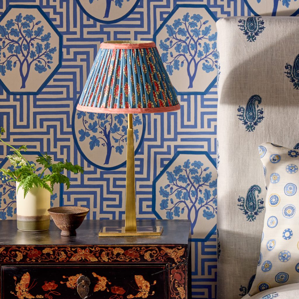 A bedside table with a brass and blue silk lamp on it, against a traditional Chinese motif blue wallpaper 