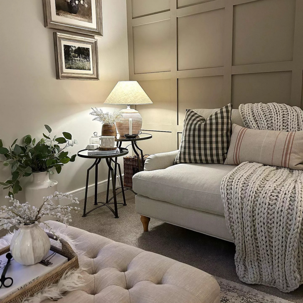 A cosy elegant living room with wood panelling, a cream sofa with chunky knitted throw, ottoman and soft lighting from a table lamp
