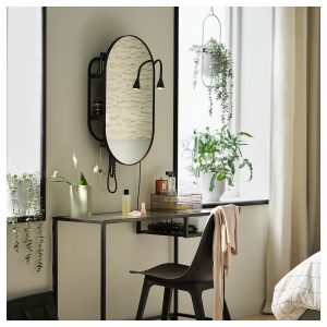 Compact wall-mounted vanity with an oval mirror, built-in shelving, and slim black frame, paired with a modern chair and surrounded by indoor plants, creating a stylish and space-efficient solution for small bedrooms.