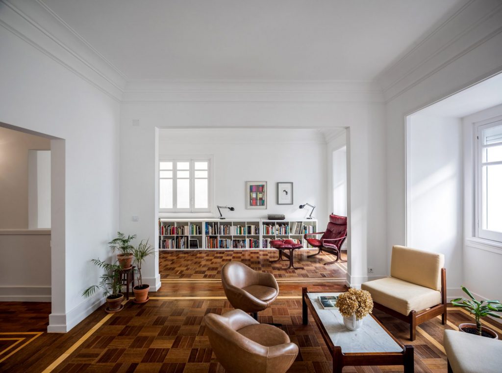 Bright living space with high ceilings, white walls, and parquet flooring, featuring mid-century furniture, indoor plants, and a built-in bookcase beneath tall windows, illustrating how morning light and architectural details enhance character and influence interior colour choices.