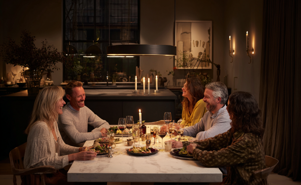Five middle-aged friends at a warmly lit dining table in a dark kitchen-dining room, enjoying an evening meal together.
